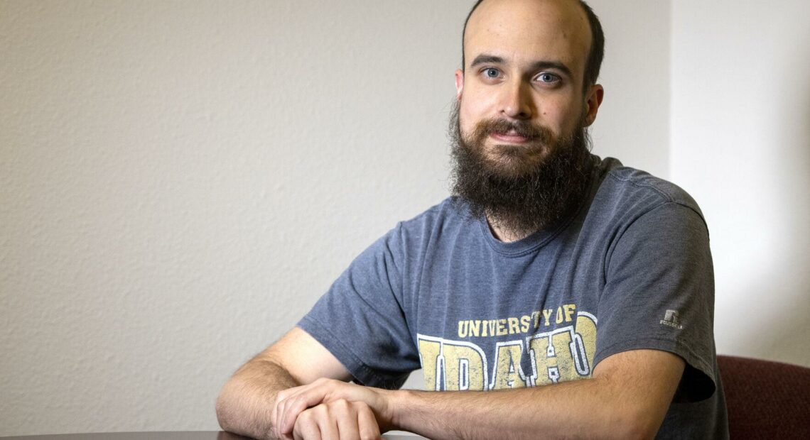 A man in a blue T-shirt with a beard sits at a table.