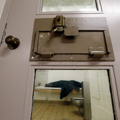 An inmate lies on a jail bed covering themself with a dark blue blanket.