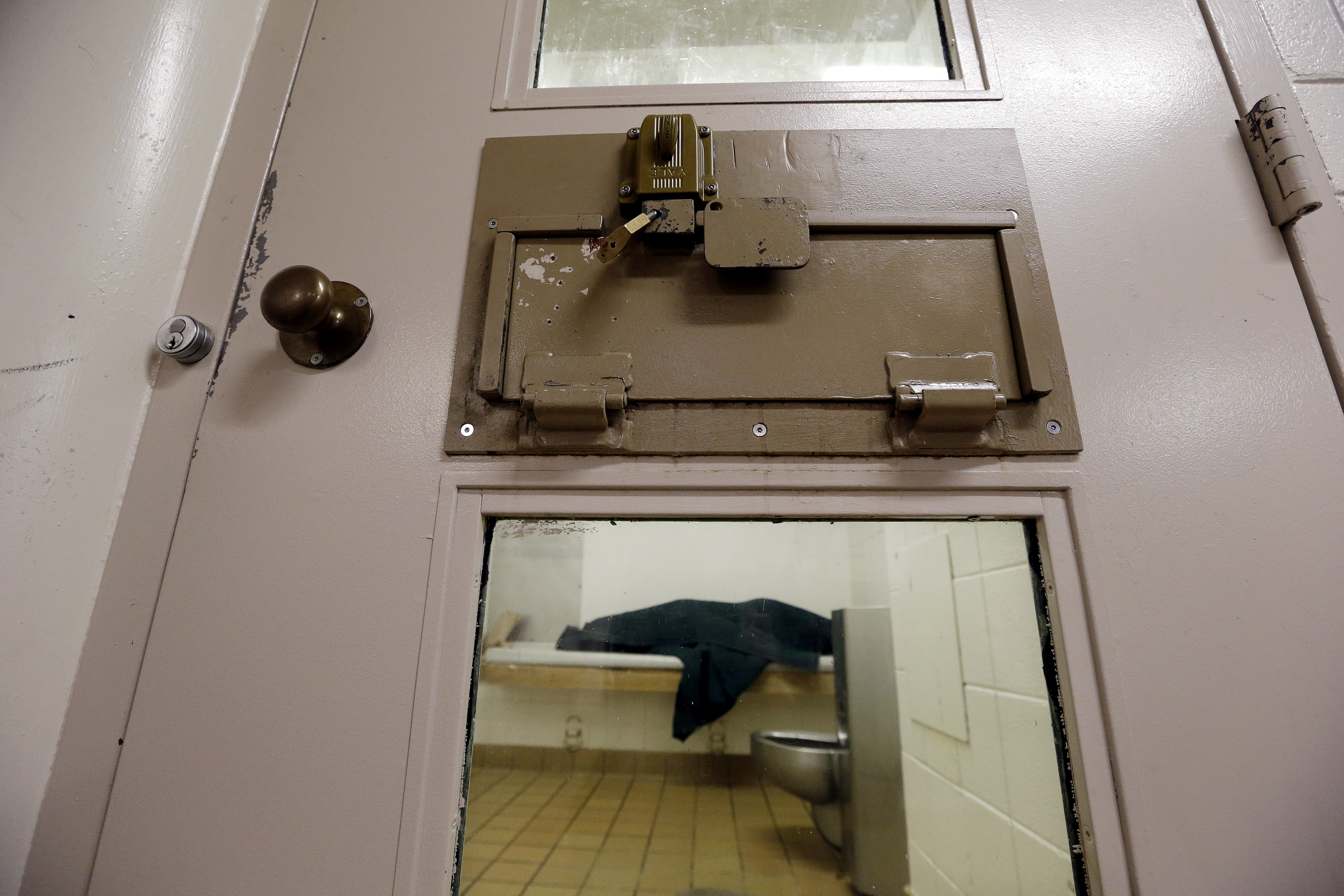 An inmate lies on a jail bed covering themself with a dark blue blanket.