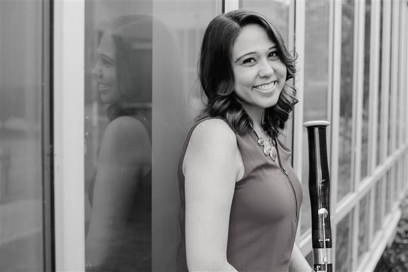 Dr. Wilson stands in front of a glass window with her bassoon. She smiles.