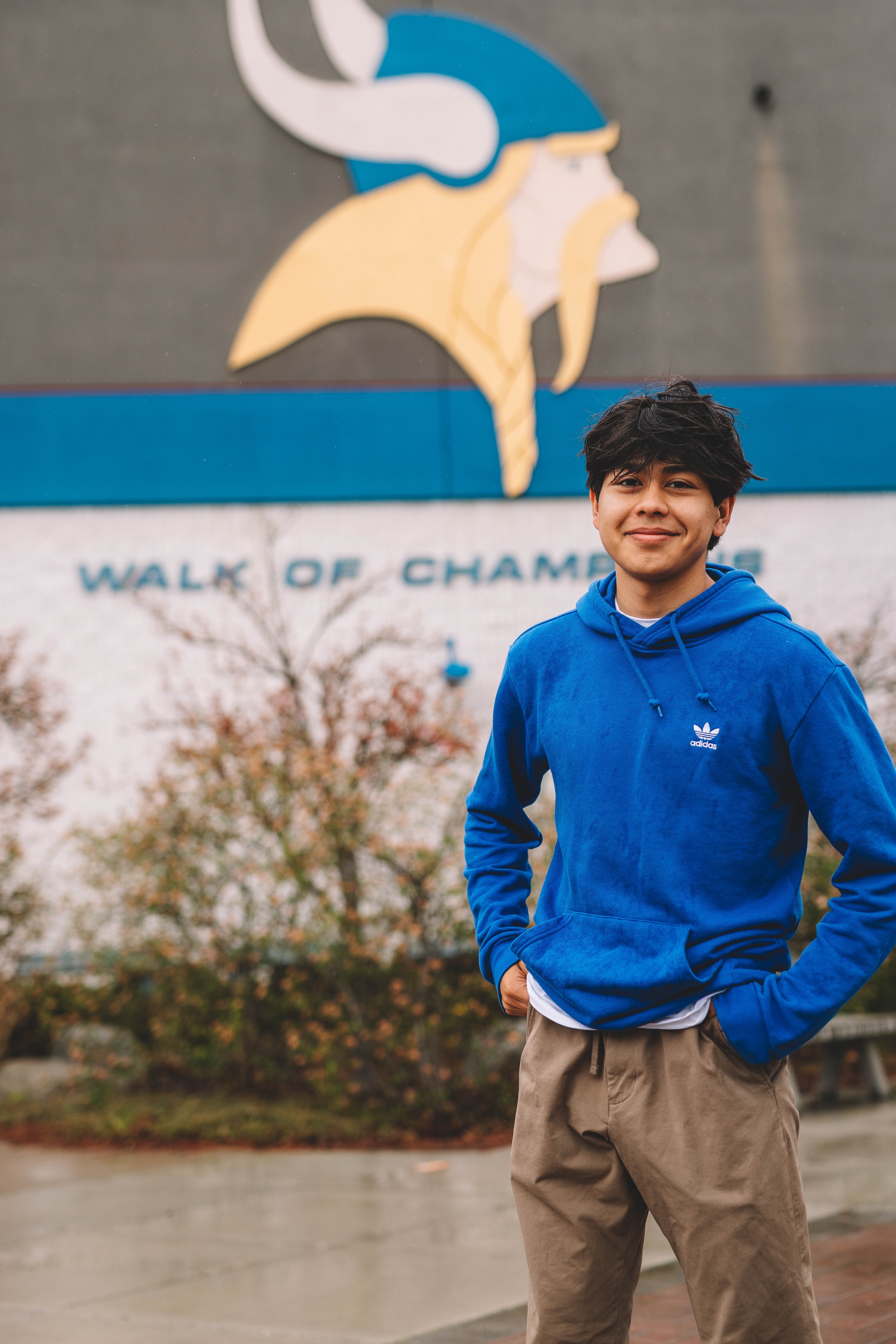 A young man of Coeur d'Alene tribal descent in a blue sweatshirt and brown pants stands in front of Coeur d'Alene high school. 