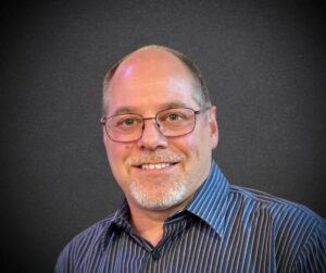 White man with glasses and a blue shirt smiling at camera 