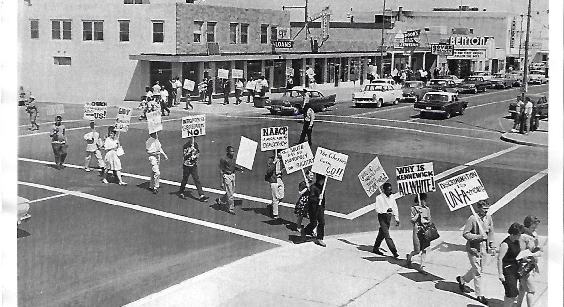 black and white photo showing people that are both Black and white marching across a street. They are holding signs protesting against discrimination.