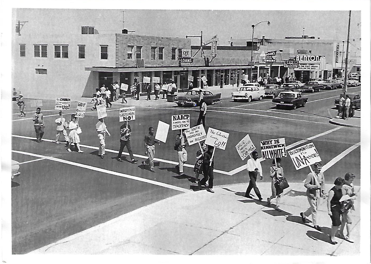 black and white photo showing people that are both Black and white marching across a street. They are holding signs protesting against discrimination.