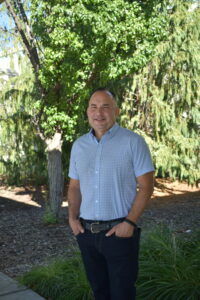 man in white shirt smiling at camera standing in front a tree