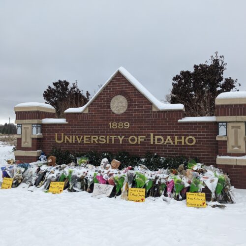 Flowers and signs are piled up on a University of Idaho sign with white snow all around the brick sign.
