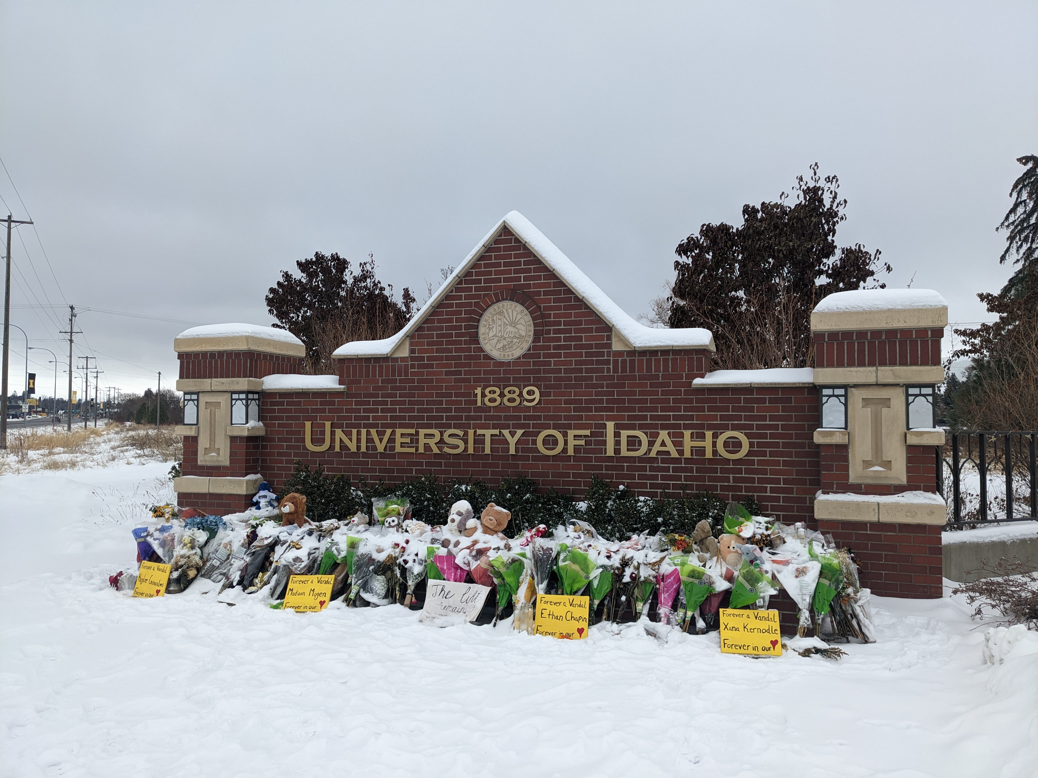 Flowers and signs are piled up on a University of Idaho sign with white snow all around the brick sign.