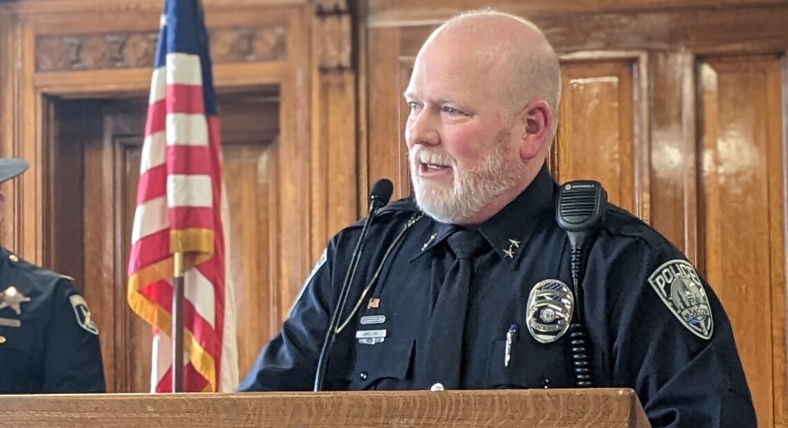 Police Chief James Fry stands before a wooden podium in his navy blue uniform in front of an American flag.