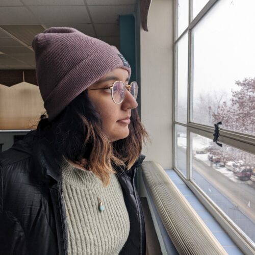 A woman with tan skin, glasses, and a purple beanie stares out the window into a snowy Moscow, Idaho.