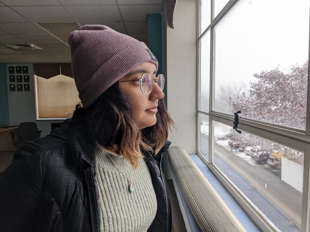 A woman with tan skin, glasses, and a purple beanie stares out the window into a snowy Moscow, Idaho.