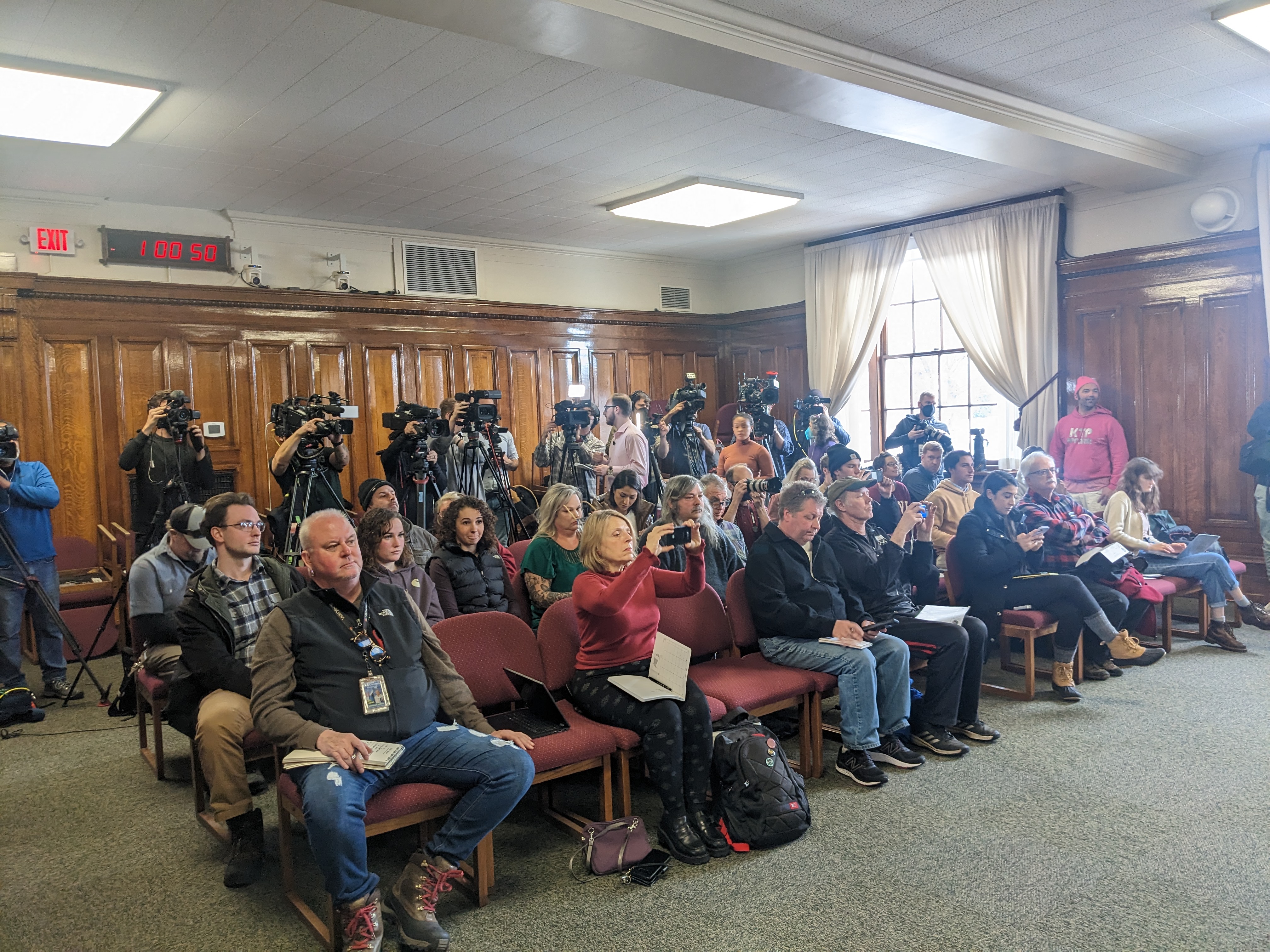 A room with green carpet is filled with people sitting in maroon folding chairs, and cameras line the back wall. 