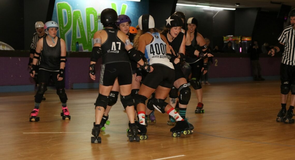 Several people on skates with pads and helmets in black and white gear play roller derby on a wooden floor.