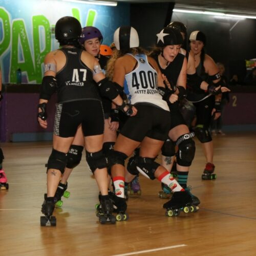 Several people on skates with pads and helmets in black and white gear play roller derby on a wooden floor.