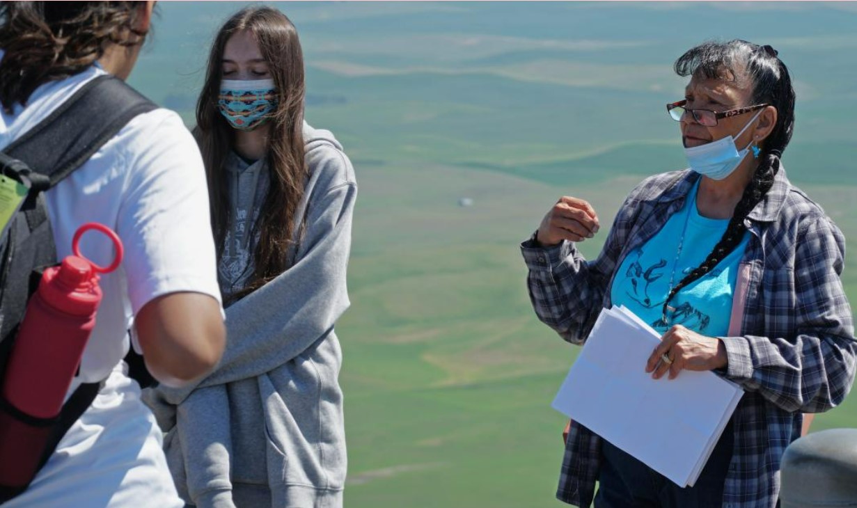 A woman with a face mask talks to students in front of green sprawling hills. 