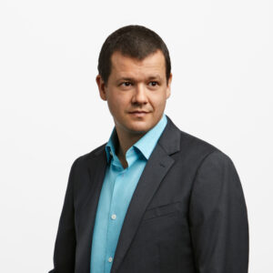 young man with dark hair looking in the distance with a dark grey jacket and light blue shirt. 