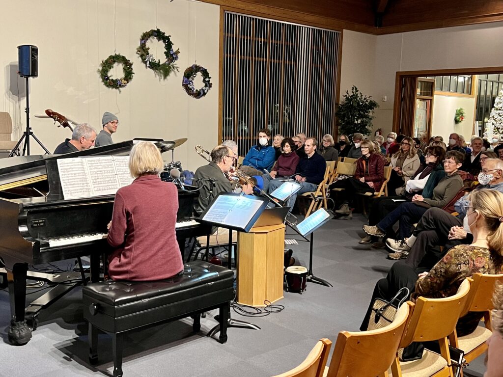 A woman in a red jacket sits at a piano. A man in a green shirt plays the trombone. Other musicians are in the background. Audience members sit in wooden chairs.