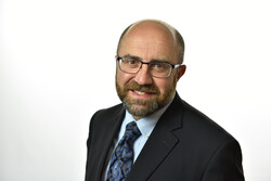 Middle aged white man with glasses and mustache and beard. Wearing a suit and smiling at the camera. 