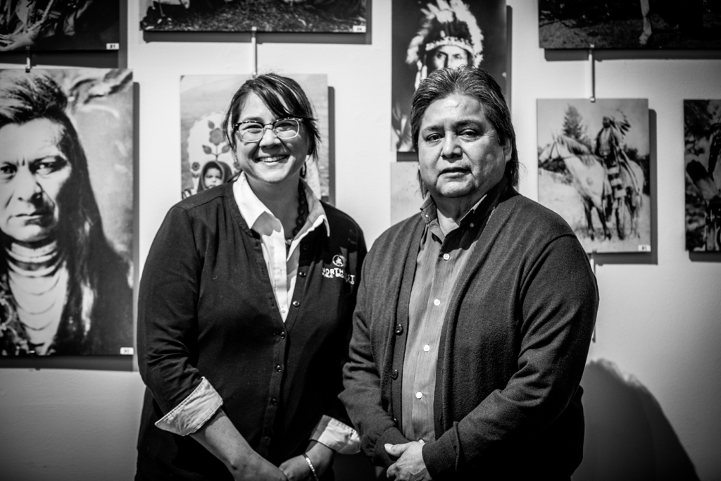 Sueann Ramella, left, and Sam Penney, right, pose together, each with their hands clasped in front of them, looking directly at the camera. Behind them is a wall of Nez Perce photographs.