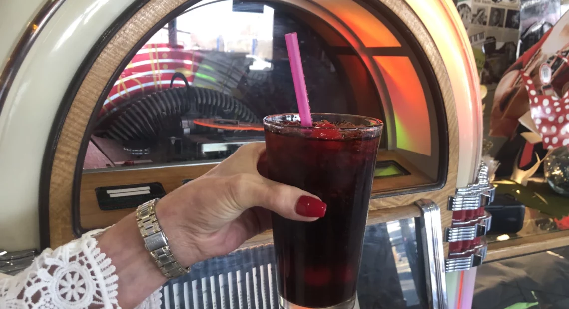 A woman with red nails holds a glass of Coca-Cola with a pink straw in front a jukebox.