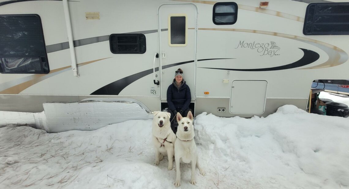 A woman wearing a black coat and winter hat sits on the steps of white RV. Two large white dogs sit in front of her on a snowy, overcast day in McCall, Idaho.