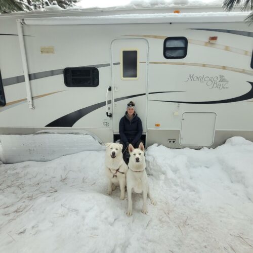 A woman wearing a black coat and winter hat sits on the steps of white RV. Two large white dogs sit in front of her on a snowy, overcast day in McCall, Idaho.