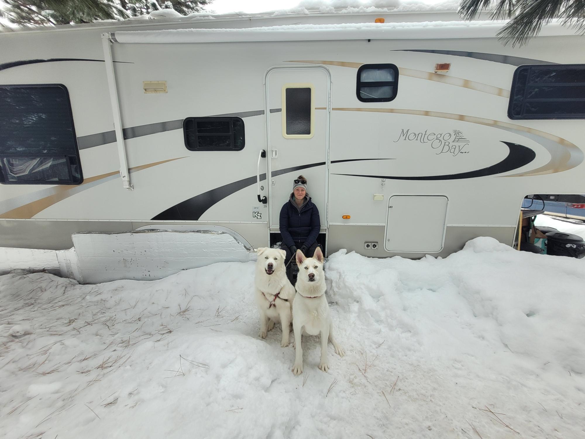A woman wearing a black coat and winter hat sits on the steps of white RV. Two large white dogs sit in front of her on a snowy, overcast day in McCall, Idaho.