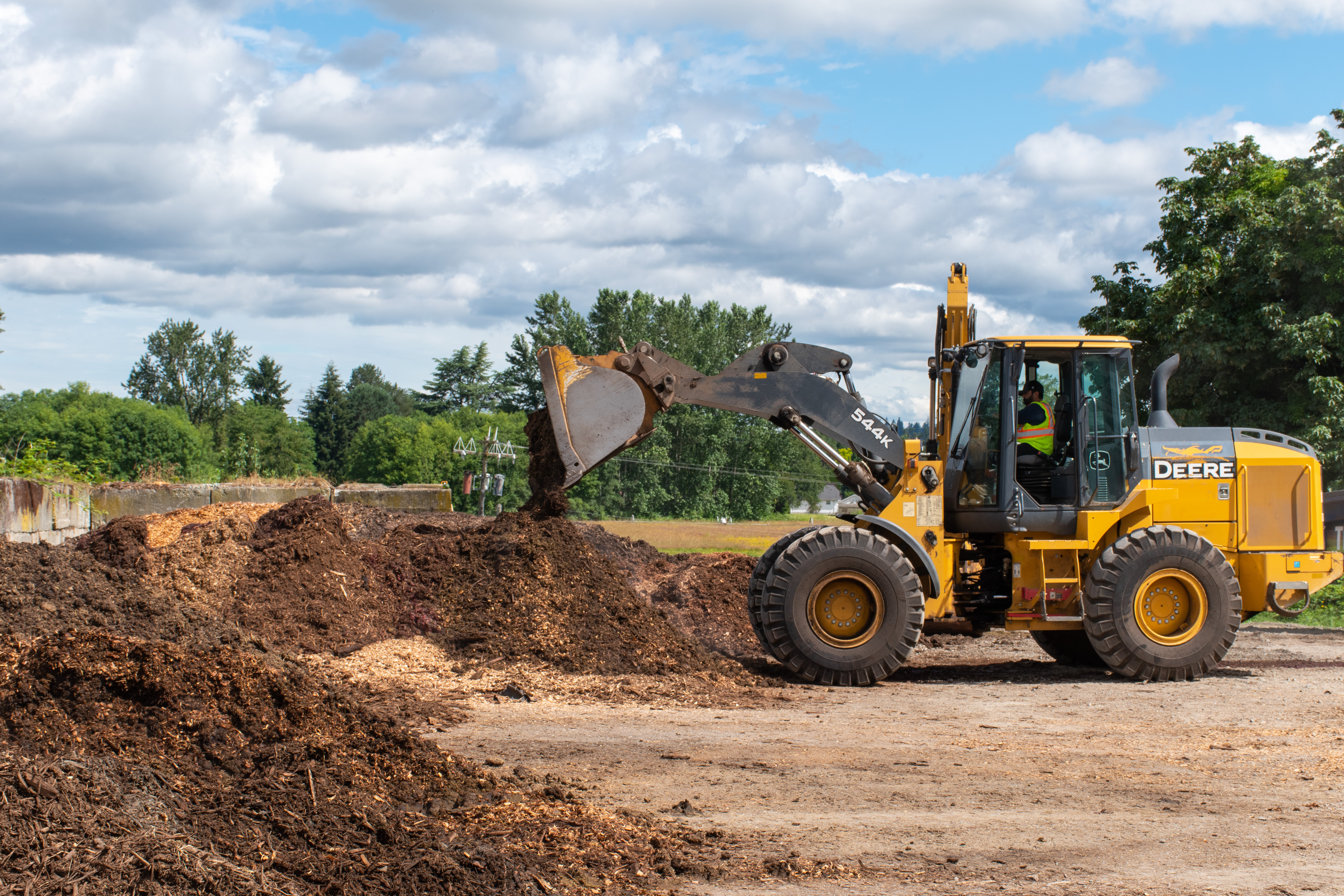 A large, yellow tractor digs into a large, brown pile of compost. Green foliage trees stand in the background beneath a blue sky with white, fluffy clouds.