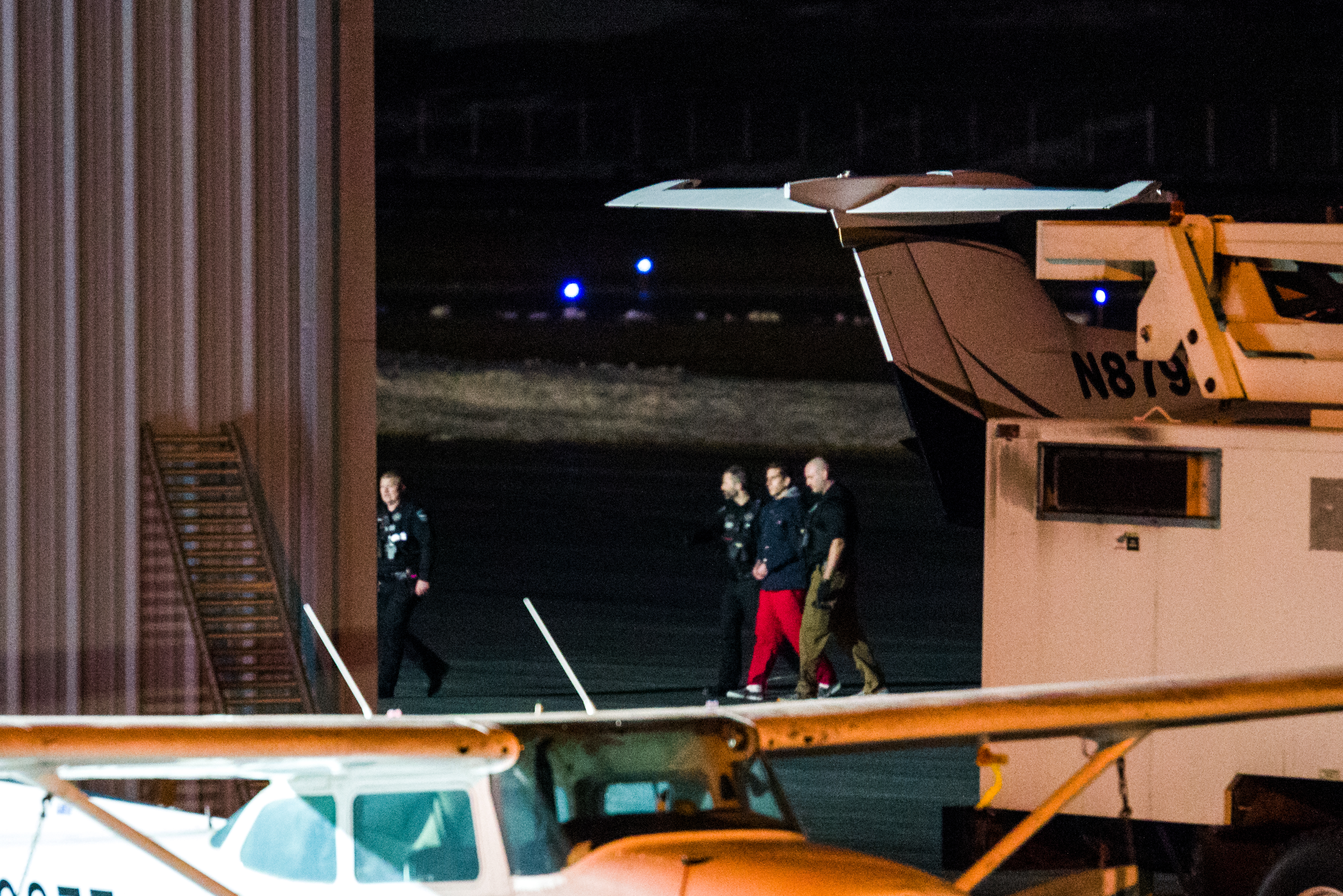 A man in an orange jumpsuit is escorted off a white plane by law enforcement officers in dark blue uniforms. The night sky hangs behind them.