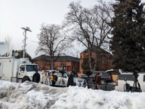 Reporters line the sidewalk in front of a pile of white snow near a white truck. Cameras are set up against the backdrop of a brick building surrounded by trees.