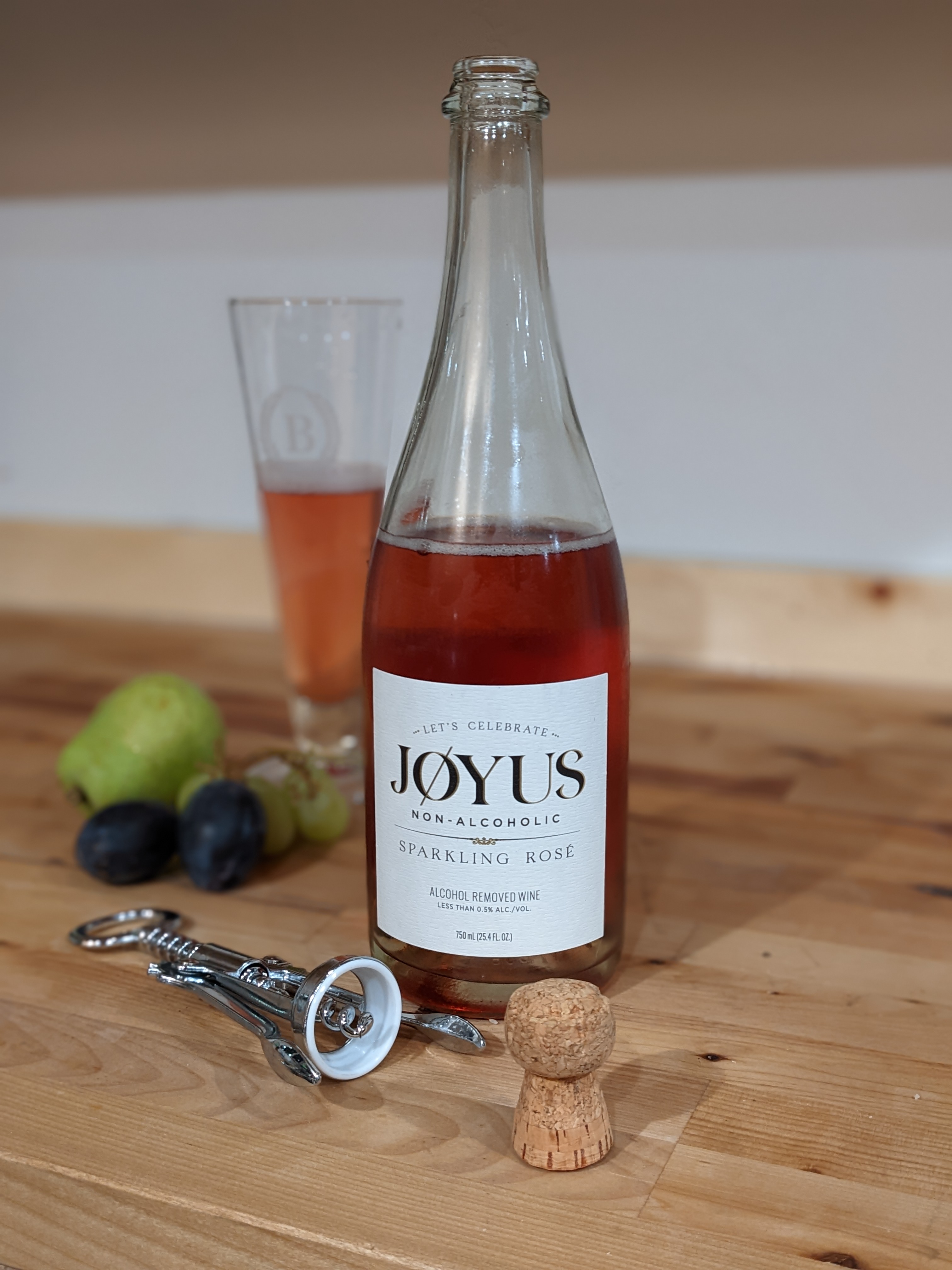 A bottle of sparkling rose wine labeled "JOYUS" sits atop a brown wood countertop next to grapes. 