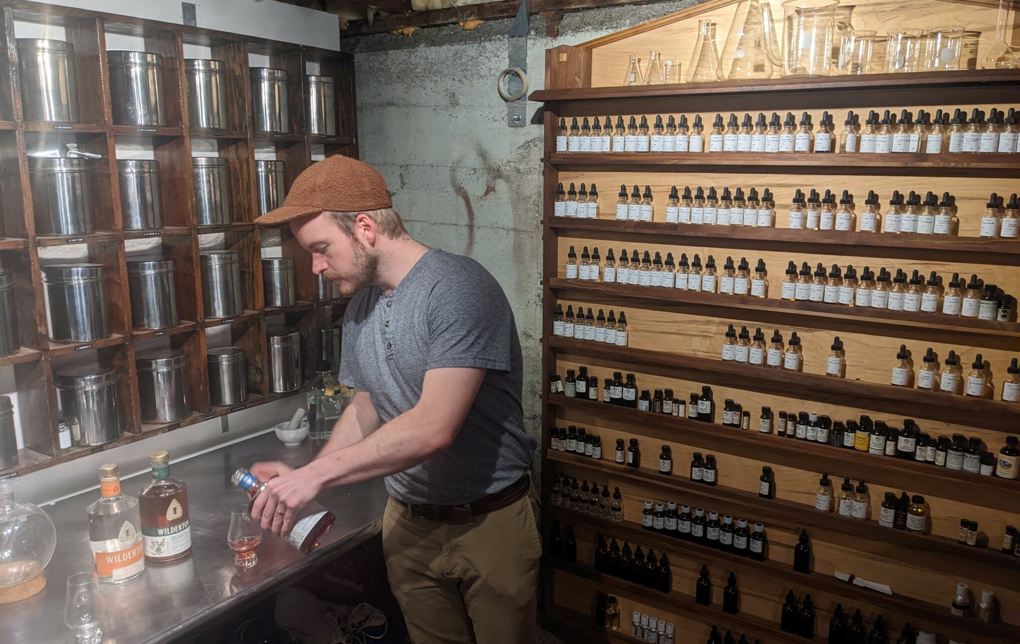 A man in a skully cap stands near a wooden wall of small shelves filled with herbs and spices. He pours non-alcoholic spirits into a clear glass.