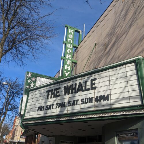 A white marquis spells out THE WHALE in black lettering on the front of an old theater with a green sign that reads "Kenworthy" against a blue sky.