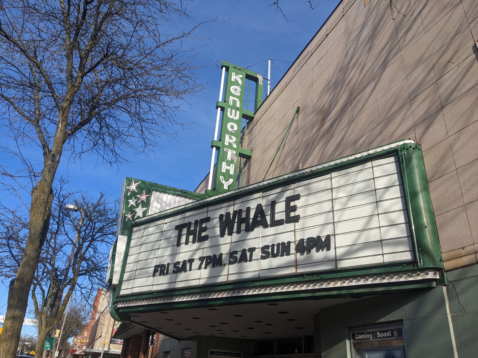 A white marquis spells out THE WHALE in black lettering on the front of an old theater with a green sign that reads "Kenworthy" against a blue sky.