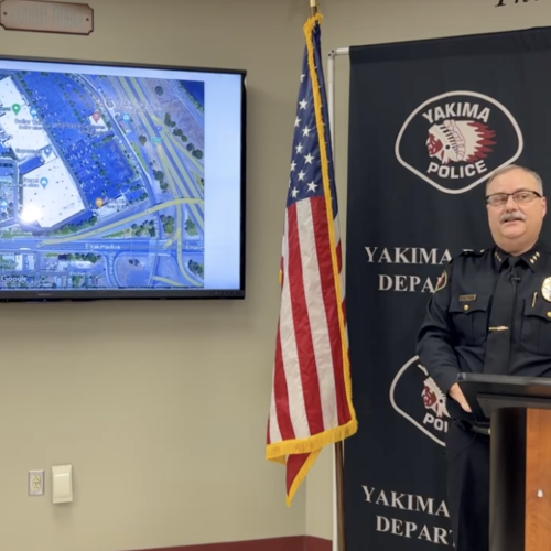 A police officer in uniform wearing glasses stands behind a podium and in front of a background reading "Yakima Police" on his left is an American flag and a computer screen showing a map of where the suspect was found dead.