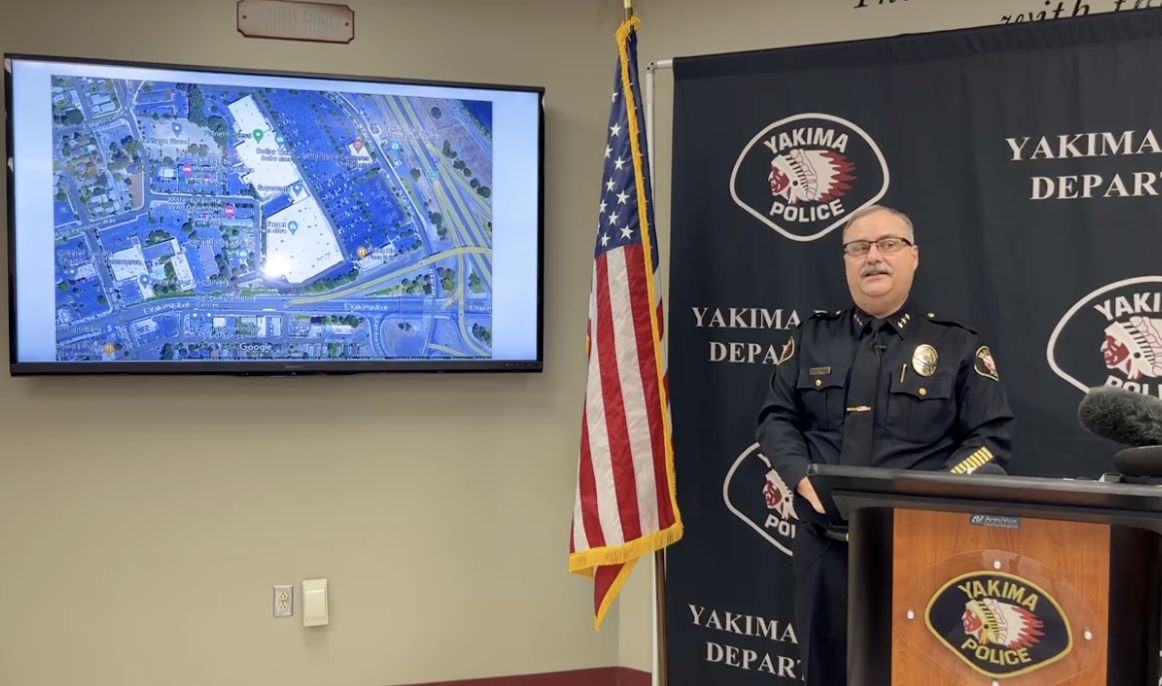 A police officer in uniform wearing glasses stands behind a podium and in front of a background reading "Yakima Police" on his left is an American flag and a computer screen showing a map of where the suspect was found dead.