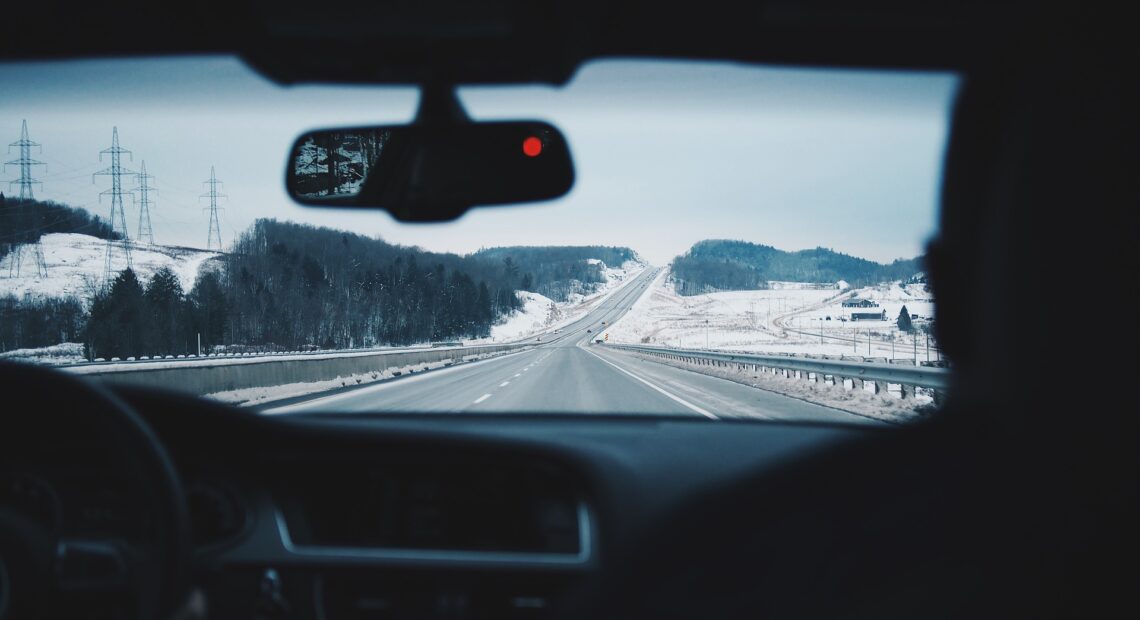 A stretch of road between snow-dusted hills and gray steel guardrails stretches in front of a dashboard and rear-view mirror of a car. The picture is taken as if the photographer were sitting in the back seat of a car.
