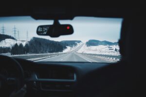 A stretch of road between snow-dusted hills and gray steel guardrails stretches in front of a dashboard and rear-view mirror of a car. The picture is taken as if the photographer were sitting in the back seat of a car.