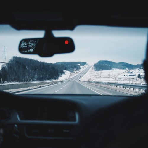 A stretch of road between snow-dusted hills and gray steel guardrails stretches in front of a dashboard and rear-view mirror of a car. The picture is taken as if the photographer were sitting in the back seat of a car.