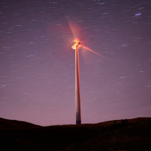 purple night sky with stars and a wind turbine with a red light in the center of the blades.