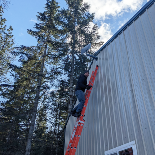 A man in jeans and a black sweatshirt climbs up an orange ladder on the side of a beige house framed by green trees and blue sky.