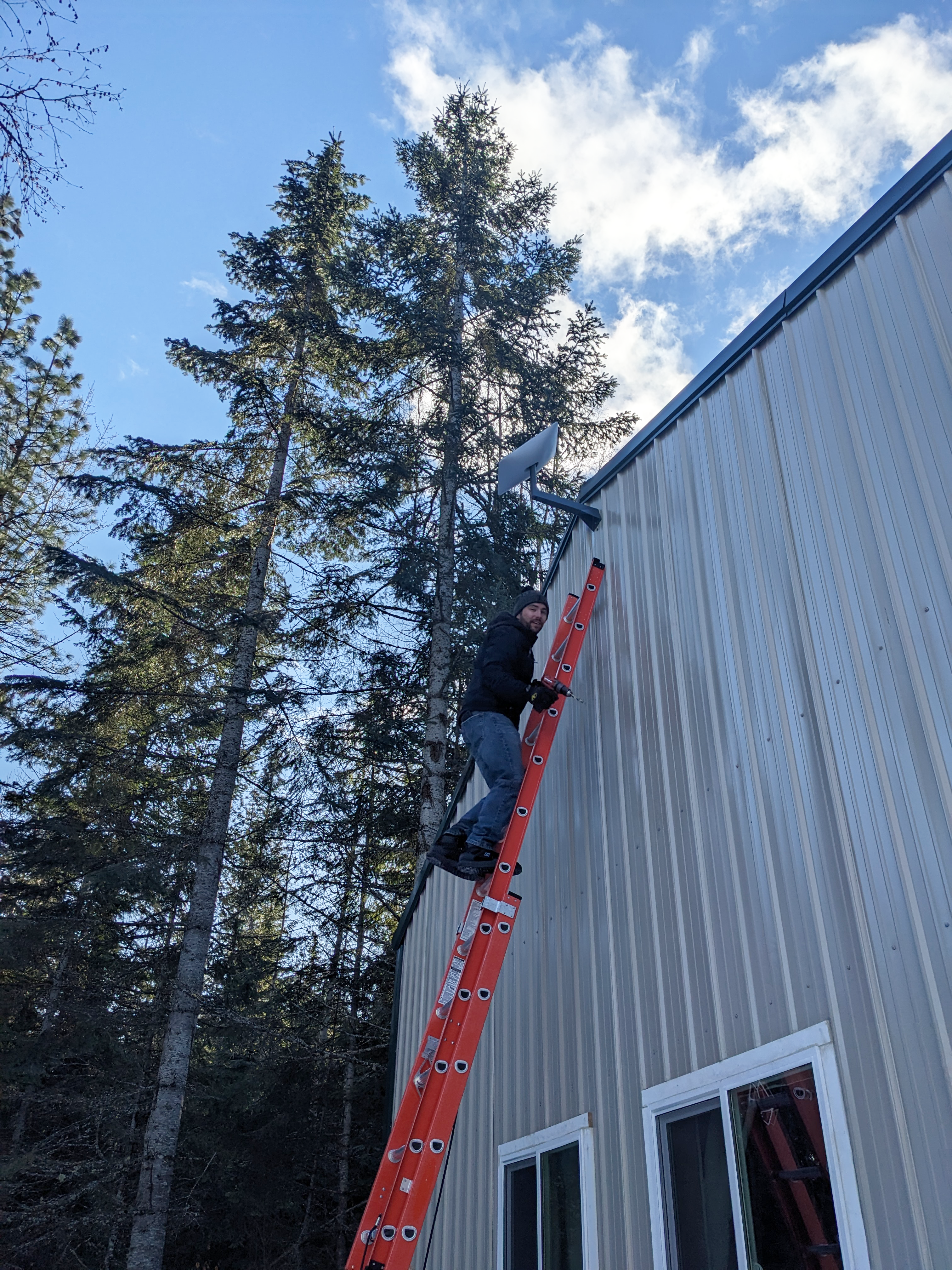 A man in jeans and a black sweatshirt climbs up an orange ladder on the side of a beige house framed by green trees and blue sky.