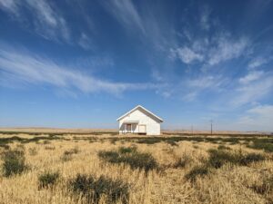 A lone, small white house surrounded by yellow sagebrush is framed against a blue sky with white, wispy clouds.
