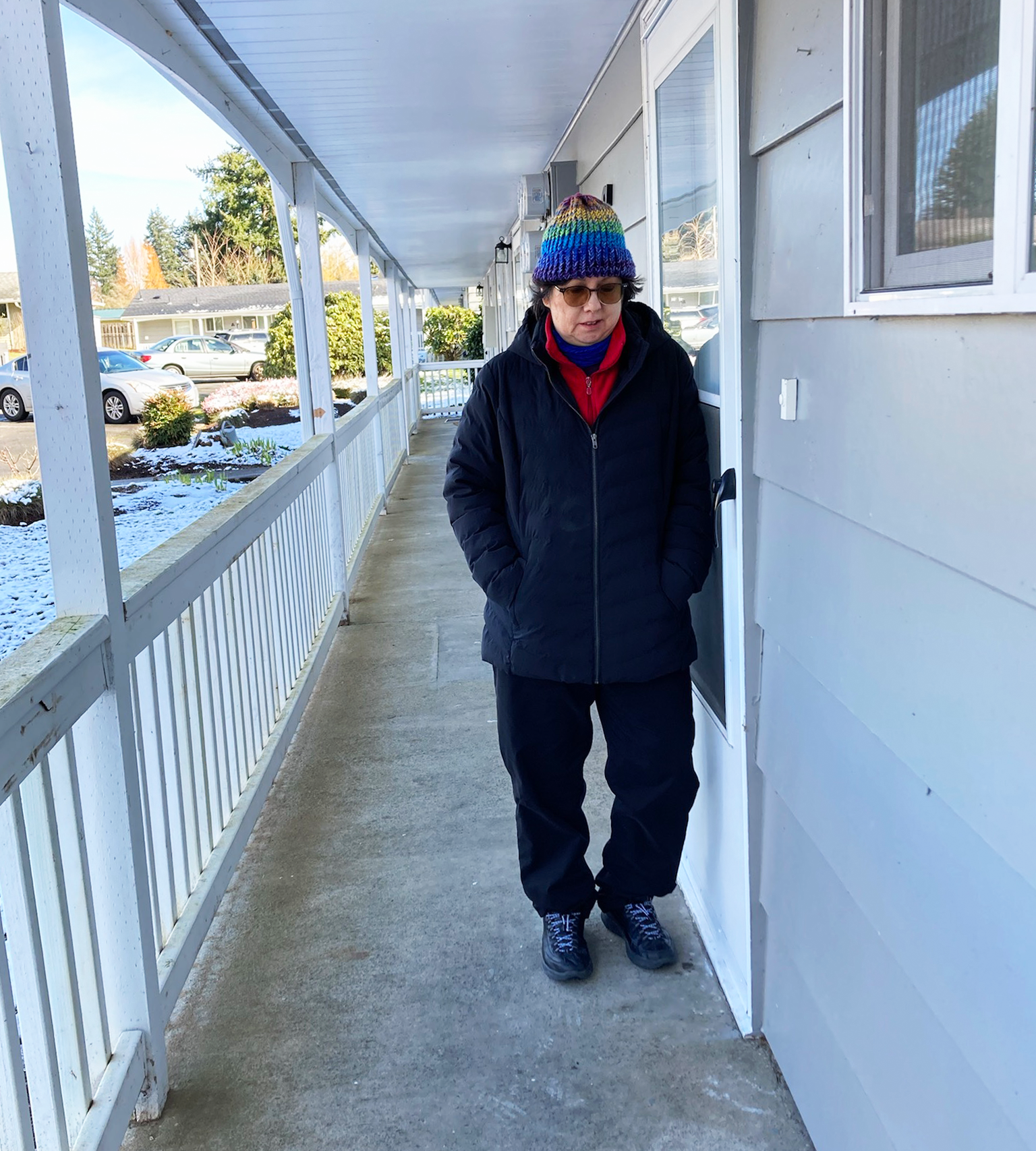 A Chinese woman in a black coat and pants and red scarf stands beside a grey apartment building. 