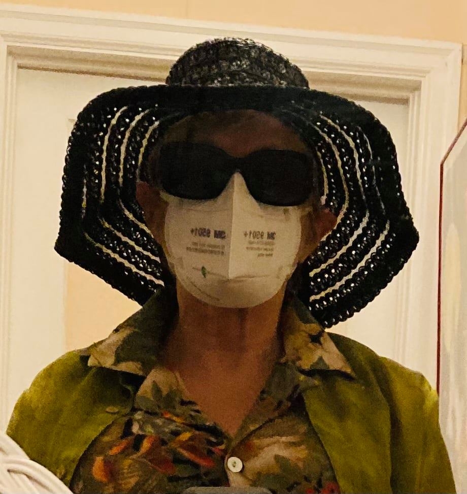 A woman stands in front of a white door frame indoors. She wears a large, floppy black hat, black sun glasses and a white, medical face mask. She wears a light green cardigan over a green flower-print button up shirt.