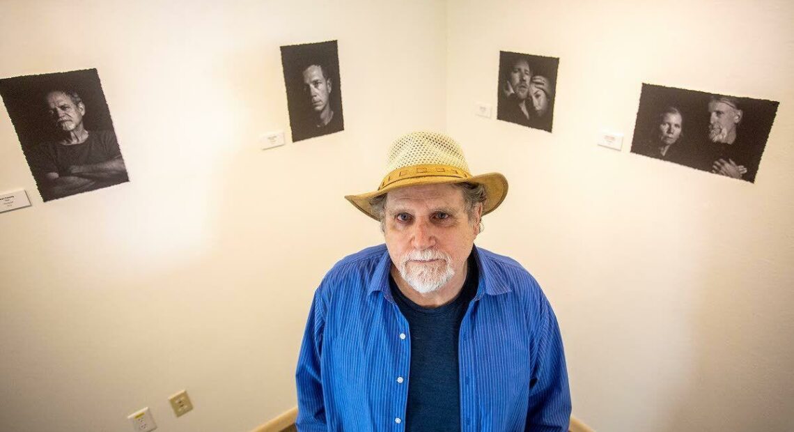 A man in a blue long-sleeved shirt over a dark blue t-shirt wearing a straw hat looks up toward the camera. The man has a short, white beard and stands in front of two walls painted off-white displaying black and white photographs.