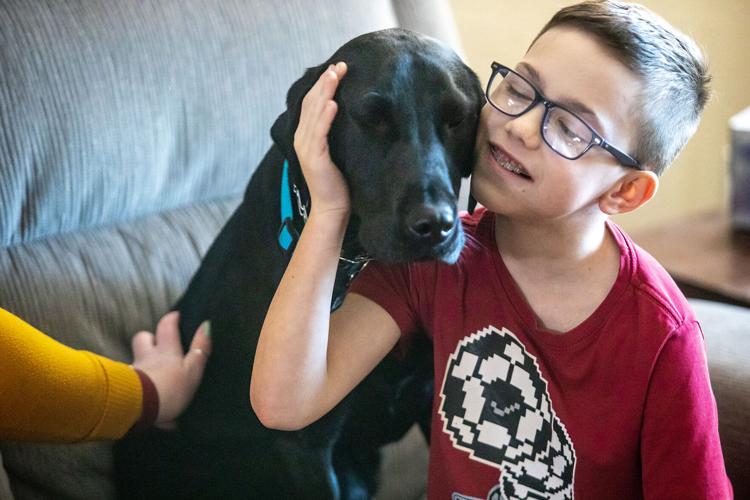 A young boy with blue-rimmed rectangular glasses closes his eyes, his mouth open in half smile showing his braces. He tilts his cheek toward a larger, black lab dog whose eyes are half closed. The boy wears a red t-shirt with digitized soccer balls printed on it. The arm of a woman stretches from the left frame of the shot and she's petting the dog wearing a yellow sweater. In the background is the cushions of a grey couch.