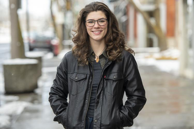 A young woman stands in a black, leather jacket with her hands in the pockets. She has long, dark, wavy hair and wears glasses with dark rims. She smiles toward the camera, in the background and out of focus is a sidewalk covered in melting snow.