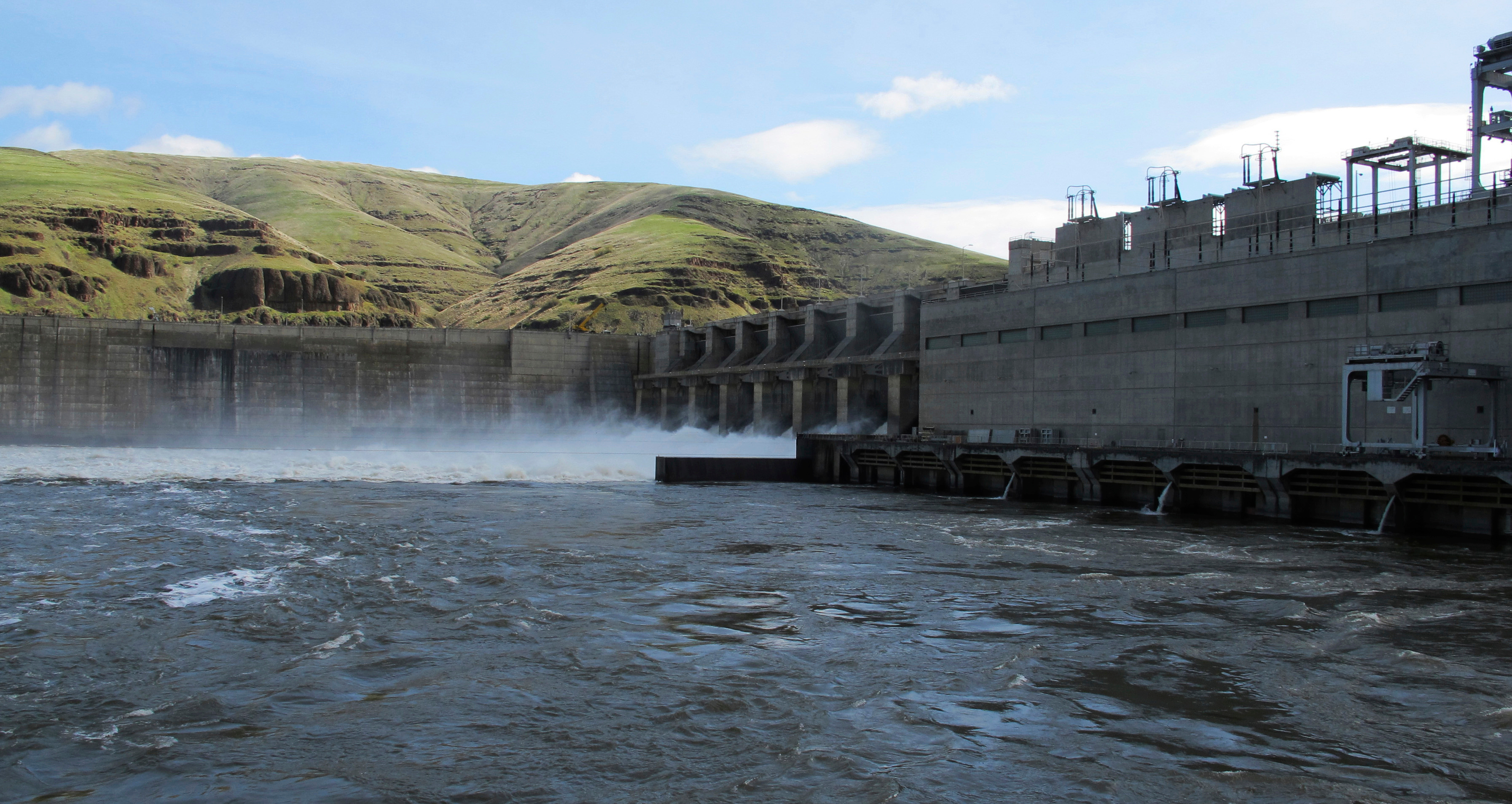 A big grey dam stretches across blue waters with mist rising from the turbines, against rolling green hills.