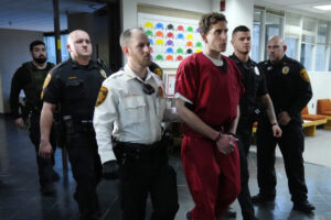 Bryan Kohberger, who appears in an orange jumpsuit with cuffs on, is flanked by uniformed police officers as they walk down a hallway.