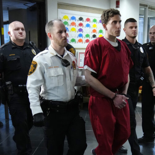 Bryan Kohberger, who appears in an orange jumpsuit with cuffs on, is flanked by uniformed police officers as they walk down a hallway.